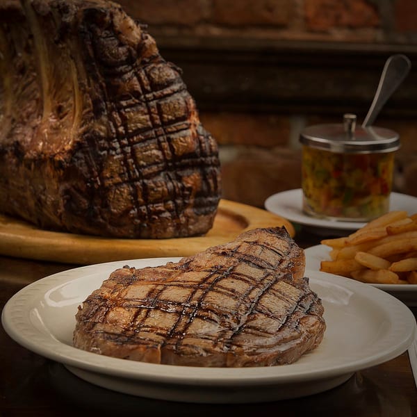 Grilled steak on a white plate with a large roast, fries, and salsa in the background.