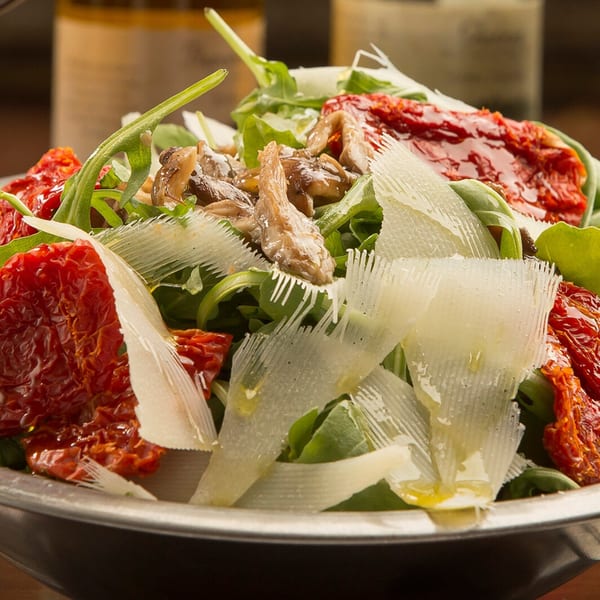 Arugula salad with sun-dried tomatoes, mushrooms, and shaved Parmesan cheese in a metal bowl.