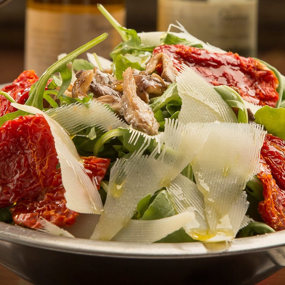 Arugula salad with sun-dried tomatoes, mushrooms, and shaved Parmesan cheese in a metal bowl.